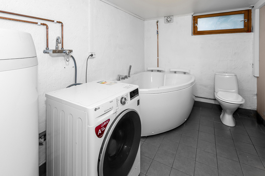 Vaskerom i kjeller med hjørnebadekar, gulvmontert toalett, VV bereder og opplegg til vaskemaskin.