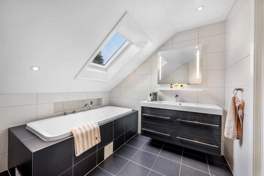 Badet har innmurt badekar, vegghengt toalett, servant i romslig innredning, veggskap og speil med innfelt lys